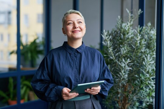 Smiling professional woman with tablet by a large window in a plant filled office for corporate profile background and marketing use