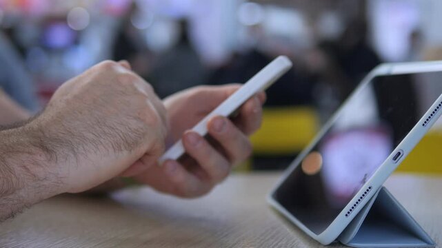 Hand holds smartphone near tablet on table. Blurred background suggests public space. Focus on digital interaction and multitasking. Suitable for tech, education, or work-from-anywhere themes