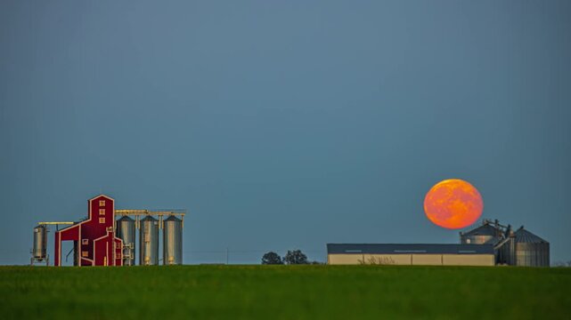 View of a large red moon rising behind farm buildings and metal grain silos in a rural landscape in Latvia.