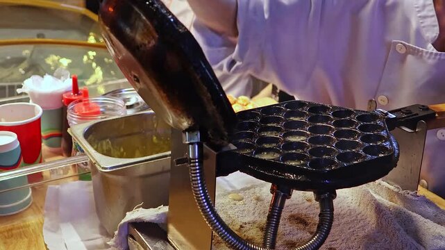 Street Food Vendor Pouring Batter Into Bubble Waffle Iron at Night Market. Close Up of Fresh Dessert Preparation, Traditional Egg Waffle Cooking Process, Outdoor Food Stall