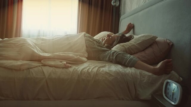 woman streching in bed waking up on sunny morning. lazy happy girl stretches ignores alarm clock continues lying sleeping enjoying weekend healthy sleep. sun light window cozy home room modern bedroom