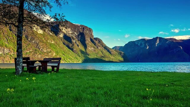 View of a wooden picnic table on a grassy bank overlooking the Aurlandsfjord with steep mountains in the background in Flam, Vestland, Norway.