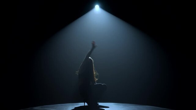 Silhouette of a female dancer performing under a single dramatic overhead spotlight on a dark stage. Concept of dance, performance, nightlife and art, fitness, fashion and entertainment advertising.