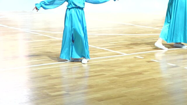 Female dancers in vibrant turquoise dresses and white shoes gracefully execute choreography across a polished wooden floor.