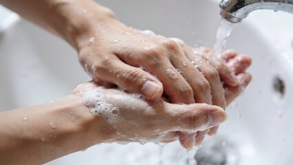 Fototapeta premium Washing adult hands under running chrome faucet at white sink basin, with soap suds and droplets