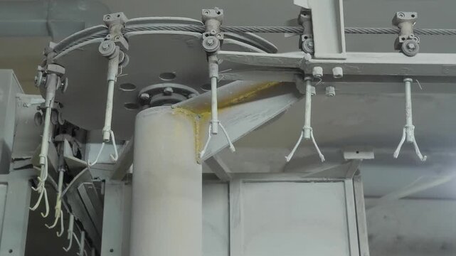Empty metal hooks on an overhead industrial conveyor line move slowly through a powder coating booth in a paint shop.