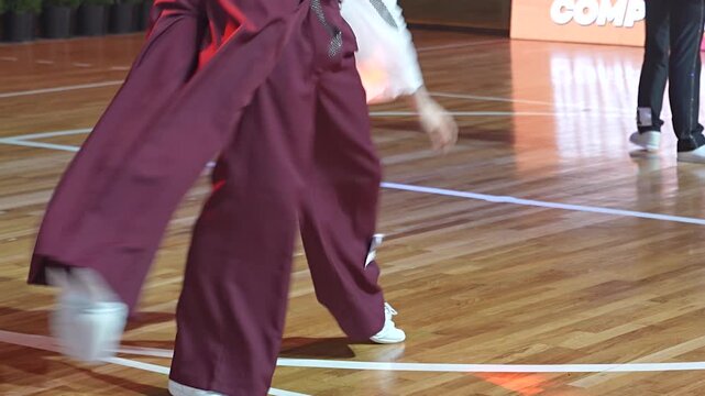 Two performers wearing loose burgundy pants and white dance shoes execute synchronized footwork across a polished wooden stage floor.