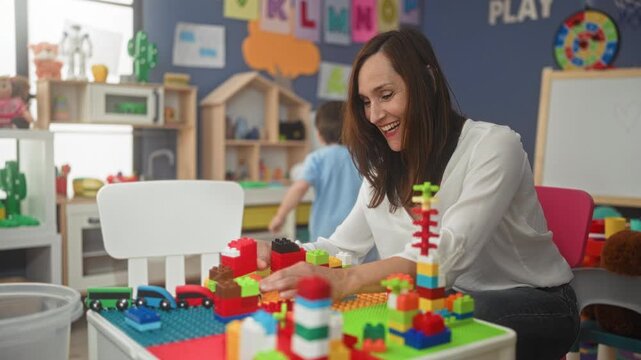 Woman reaches hand arranging toy blocks while little boy holds a toy truck in preschool building; learning play nurturing.
