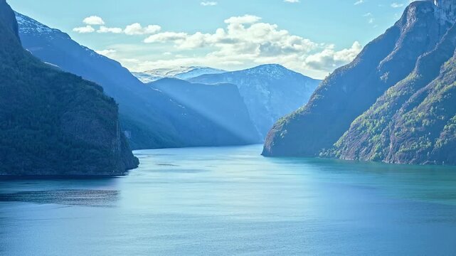 View of the deep blue waters of Aurlandsfjord flanked by towering snow-capped mountains under a bright sky in Flam, Vestland, Norway.