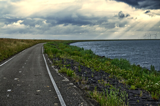 Curved dike road offering view of wind turbines generating clean energy over large lake under cloudy sky