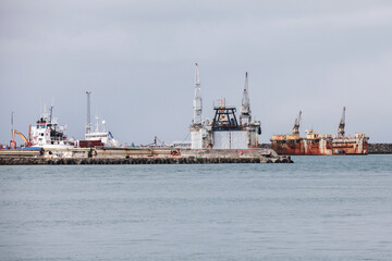 Fototapeta premium Industrial harbor scene with docked ships, rusted cargo vessels and heavy cranes along a cloudy coastal pier, conveying maritime industry, weathered infrastructure and a quiet seafront atmosphere