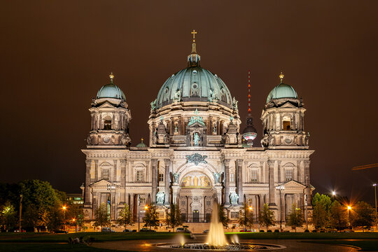Night view of the illuminated Berliner Dom (Berlin Cathedral) with its green domes, an illuminated fountain in the foreground, the Fernsehturm visible behind the cathedral, and parked cars along the s