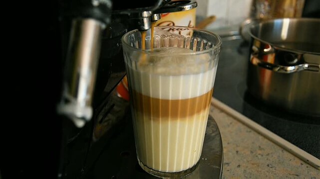 Clear ribbed glass holds thick white milk as dark coffee pours steadily from metal spout forming layered drink