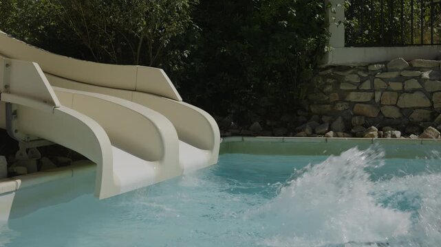 Girl slides down a white water slide into a swimming pool, splashing water, slow motion, wearing swimming goggles, outdoor pool surrounded by green bushes and a stone wall
