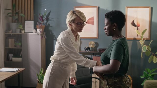 Middle aged Caucasian female therapist talking to young adult Black female soldier patient, touching her arm to offer support. Footage is ideal for mental health services and clinic commercials