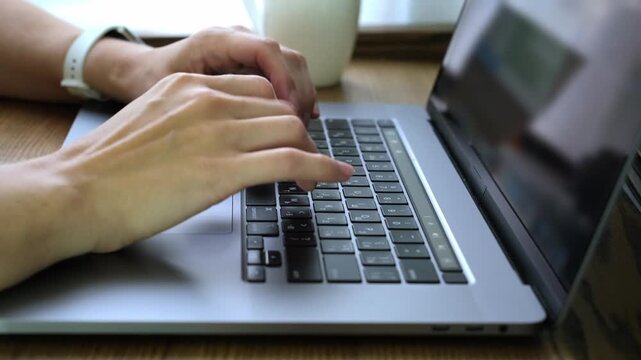 Side view close up of female hands typing on a laptop keyboard while sitting at a wooden table working online at home or in an office.