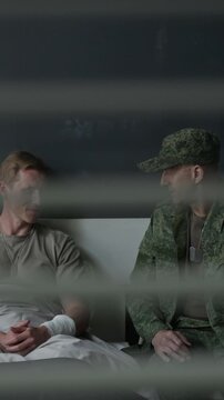 Vertical shot of young adult Caucasian soldier with bandaged arm lying in hospital bed and talking to middle-aged Caucasian officer. Footage for veteran rehabilitation stories