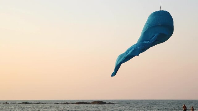 A blue fabric windsock flutters over the sea at sunset over the coastal horizon, slow motion