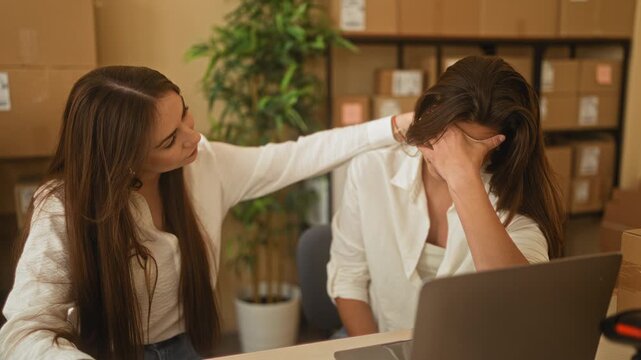 Woman comforts colleague with hand to head over laptop in small business building; empathy support teamwork.