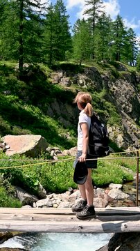 Young woman with a backpack enjoying the pristine mountain landscape while standing on a small wooden bridge crossing a clear stream in the middle of a lush forest on a beautiful sunny day