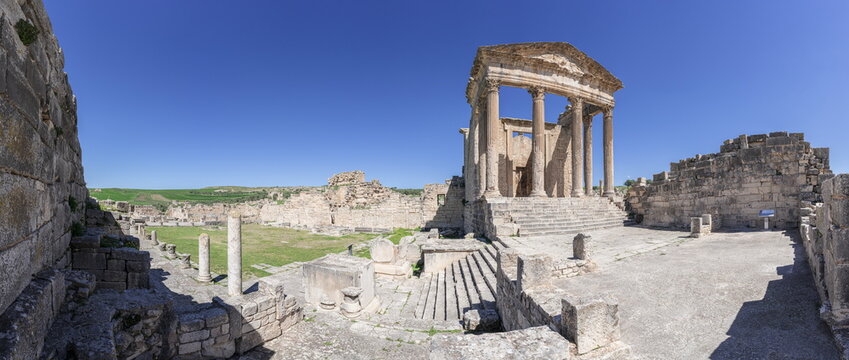 View of the Capitol temple at the Dougga Archaeological Site with ancient stone columns, ruins, and green fields under a clear blue sky in Teboursouk, Beja Governorate, Tunisia.