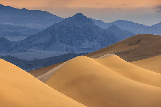 View of the golden sand dunes of Death Valley National Park with rugged mountains in the background under a warm sunset sky in the United States.