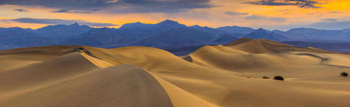 View of the sand dunes at Death Valley National Park under a golden sunset sky with distant mountains in the background, United States.
