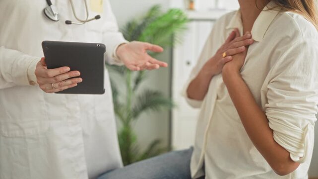 Woman patient holds hand to chest while doctor in white coat holds tablet and stethoscope visible in building; reassurance care.