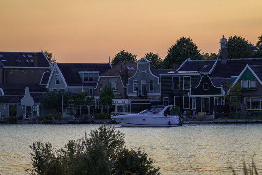View of a motorboat cruising on the Zaan river in front of traditional wooden houses at Zaanse Schans during a warm sunset in Koog aan de Zaan, North Holland, Netherlands.