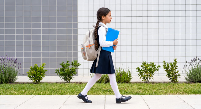 A side view of a young caucasian schoolgirl with a long braid wearing a dark blue uniform and a backpack, walking outside holding a blue folder.
