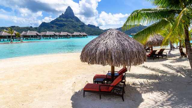 View of overwater bungalows on the turquoise lagoon with Mount Otemanu and sun loungers under a thatched umbrella on the sandy beach in Bora-Bora, Leeward Islands, French Polynesia.