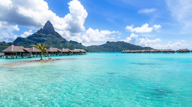 View of overwater bungalows in the turquoise lagoon with Mount Otemanu in the background under a blue sky with white clouds, Bora-Bora, Leeward Islands, French Polynesia.