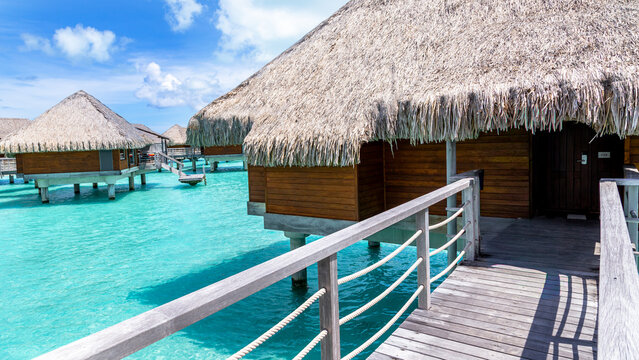 View of overwater bungalows with thatched roofs connected by wooden walkways over the turquoise lagoon in Bora-Bora, Leeward Islands, French Polynesia.
