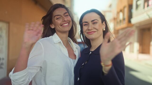 Two women smile and wave hands to camera on a sunlit narrow street with buildings and balconies, wearing casual jackets and earrings; friendship farewell joy.