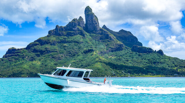 View of a white motorboat cruising across the turquoise lagoon with the lush green slopes of Mount Otemanu in the background Bora-Bora, Leeward Islands, French Polynesia.