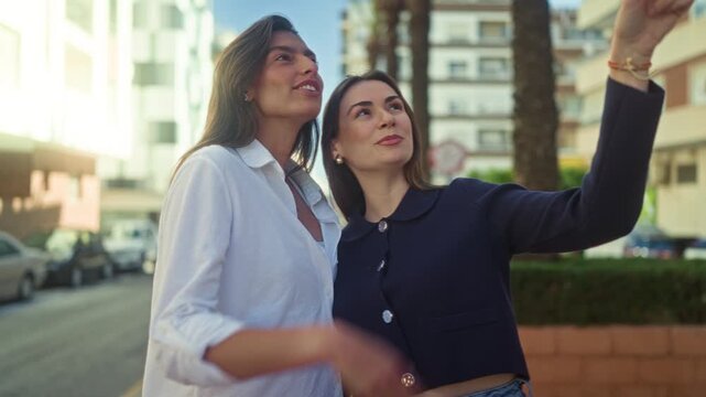 Two women pointing and smiling on a sunlit street by buildings and palm trees, one with earrings and a navy jacket; friendship joy.