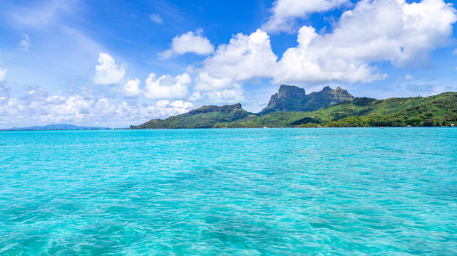 View of the turquoise lagoon of Bora-Bora with Mount Otemanu rising in the background under a bright blue sky with white clouds in Bora-Bora, Leeward Islands, French Polynesia.