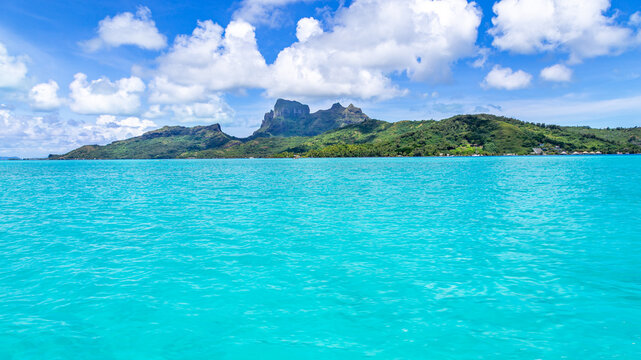 View of Mount Otemanu rising above the turquoise lagoon with lush green slopes and a bright blue sky with scattered clouds Bora-Bora, Leeward Islands, French Polynesia.