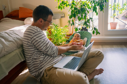 Father holding his baby's feet while sitting on the floor with a laptop, managing his professional tasks and family life simultaneously in a bright home environment