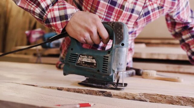 Craftsman cutting live edge wooden plank with jigsaw, guiding saw along timber edge during woodworking project on workbench inside carpentry workshop