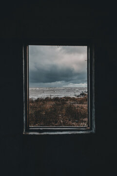 View of the stormy sea and rocky coastline of Vormsi island framed by a dark interior window, with dramatic dark clouds and dry coastal vegetation in Laane County, Estonia.