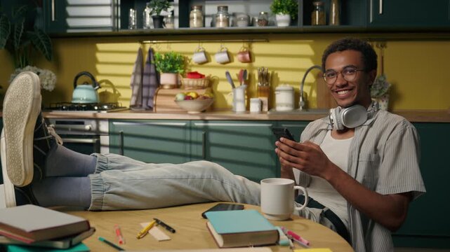 Smiling African American man scrolling mobile phone at home kitchen with legs on table young student freelancer relaxed during study break texting shopping online ordering delivery internet browsing