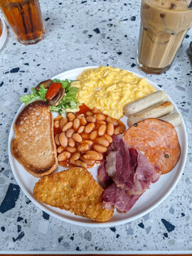 Close-up of a traditional full English breakfast plate featuring scrambled eggs, baked beans, sausages, toast, and a hash brown on a terrazzo table.