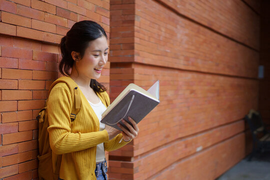 Young asian female student with backpack smiling while holding a book and a takeaway coffee cup, walking outside a modern building university campus