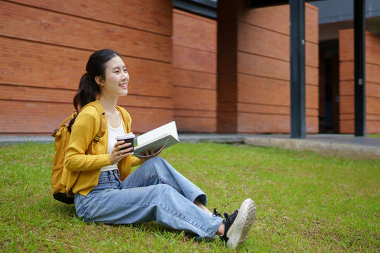 Young Asian woman student enjoying outdoor study reading a book and drinking coffee in a university campus park, highlighting education and remote learning lifestyle