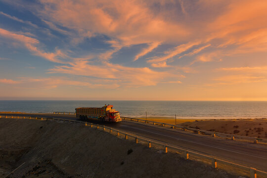 View of a decorated truck driving along the Makran Coastal Highway during a vibrant sunset with the sea and desert landscape in Balochistan, Pakistan.