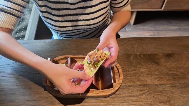 woman holding delicious taco with meat and salsa at wooden restaurant table. fresh taco sits in metal holder on serving board. concept of enjoying tasty taco meal in cafe for lunch or dinner time