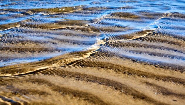 Showing wet rippled sand and thin water flowing at sandy shoreline, with sky reflections