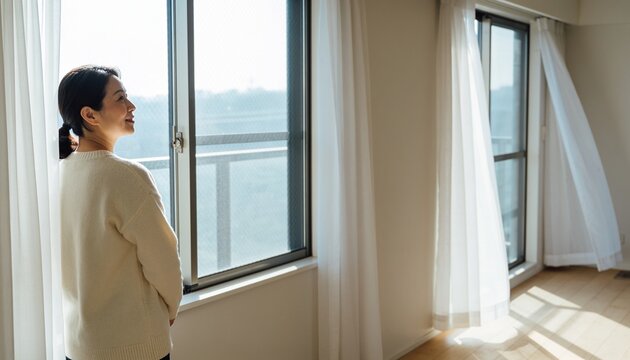 Gazing Asian woman in cream sweater dark pants looking out sliding windows at apartment, copy space