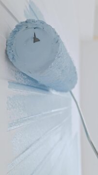 A hand uses a paint roller to apply light blue paint on an interior wall, creating smooth vertical strokes during a home renovation